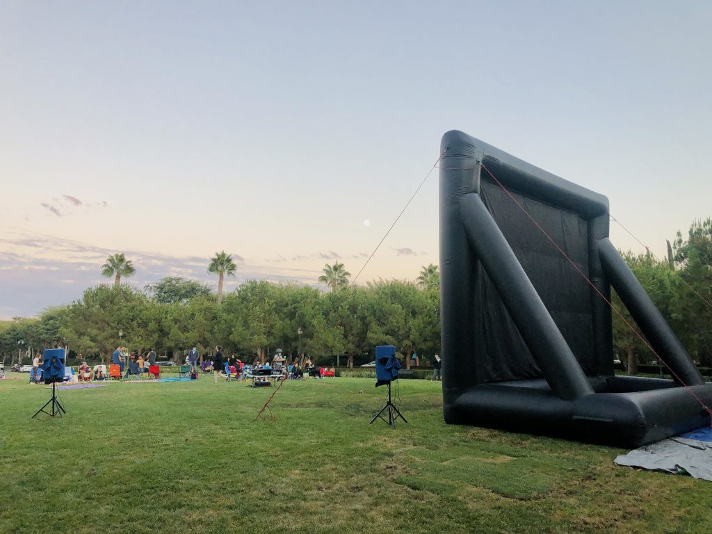 A crowd of people waiting for the outdoor movie to start when the sun sets at a local parks and recreation event.