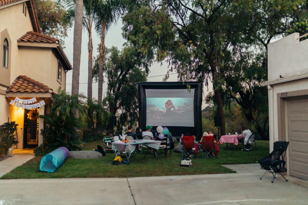 Family and friends watching The Little Mermaid on a huge FunFlicks inflatable screen in the backyard. They are celebrating a special birthday with food, party favors, and a movie night.